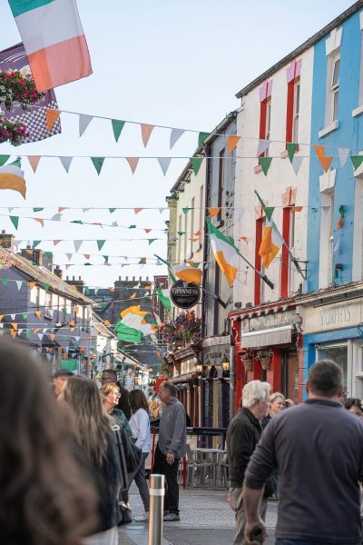 Festive street scene in Galway, Ireland, with colorful facades, Irish flags, and people walking under strings of bunting on a lively evening.