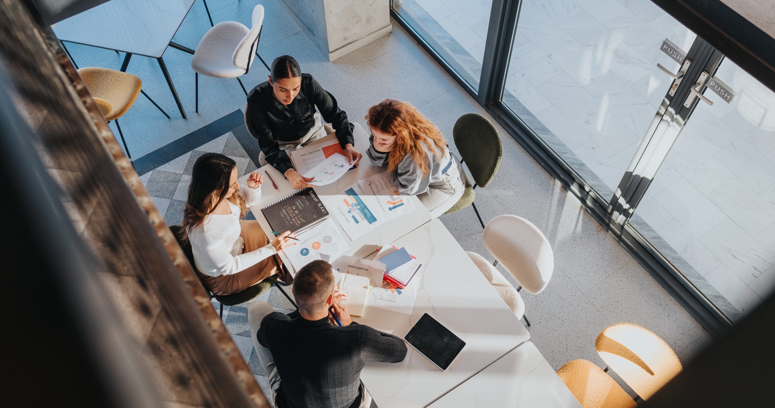 A high-angle view shows a diverse team gathered around a white table, reviewing reports and charts. Collaboration, planning, and teamwork drive a productive business meeting in a bright, open space.